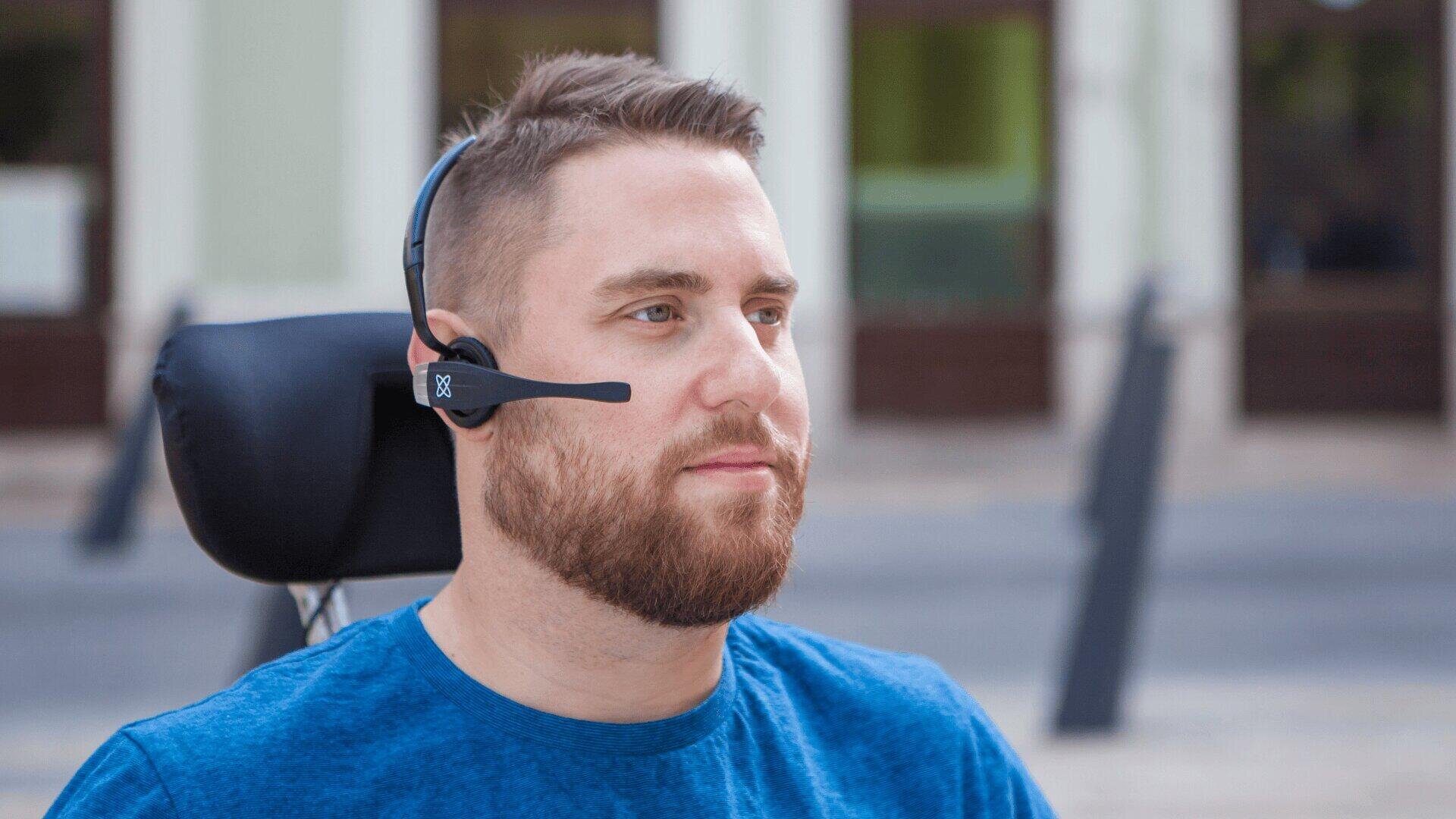 Ein bärtiger Mann mit blauem Hemd und Headset-Mikrofon sitzt in einem Rollstuhl im Freien, mit verschwommenen Gebäuden und Straßen im Hintergrund.