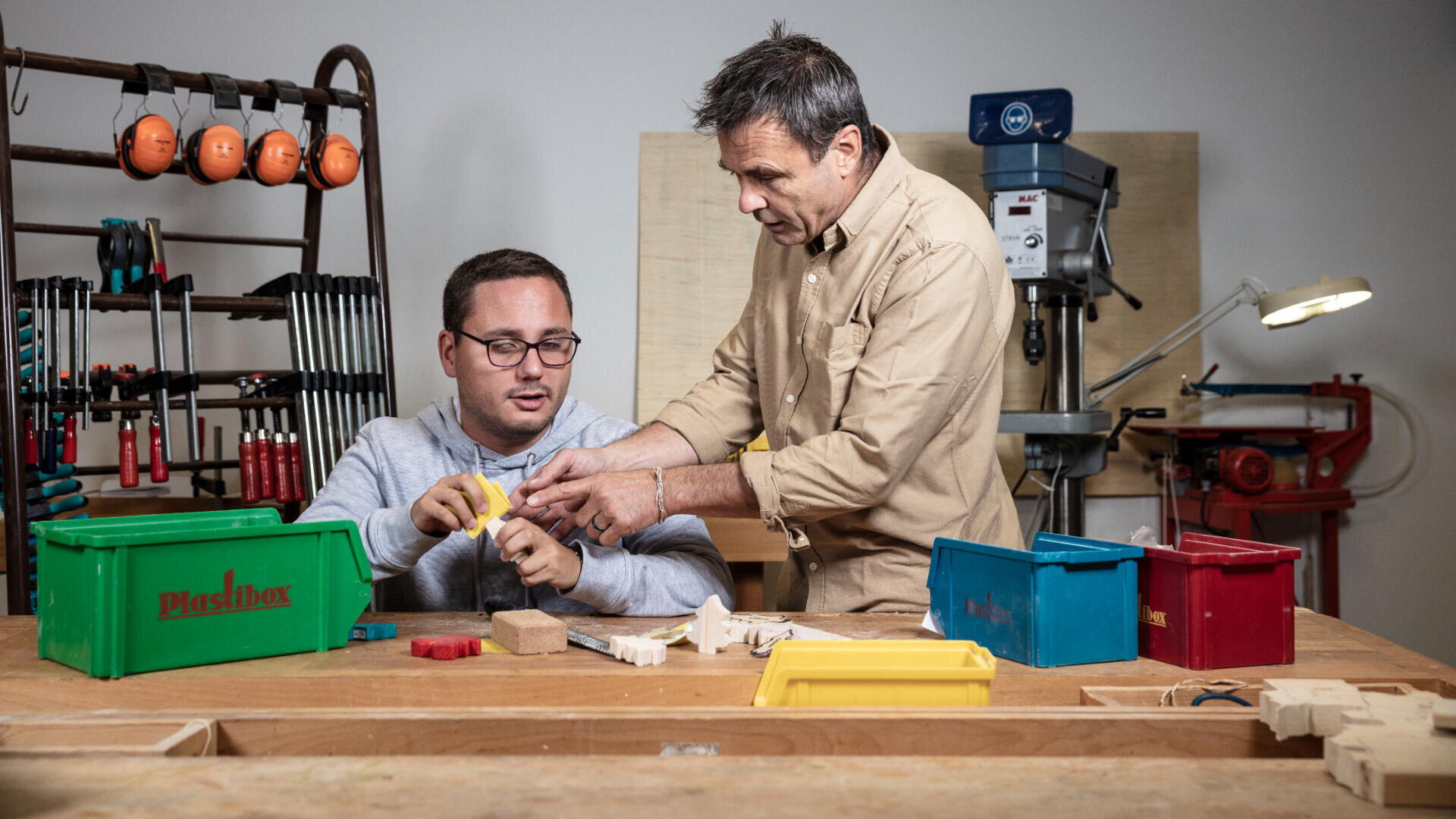 Zwei Männer arbeiten gemeinsam an einem Werkstatttisch mit verschiedenfarbigen Plastikbehältern und Holzteilen. Ein Mann sitzt und hält ein Werkzeug in der Hand, während der andere neben ihm steht und ihn anleitet. Im Hintergrund sind Werkzeuge und Maschinen zu sehen.