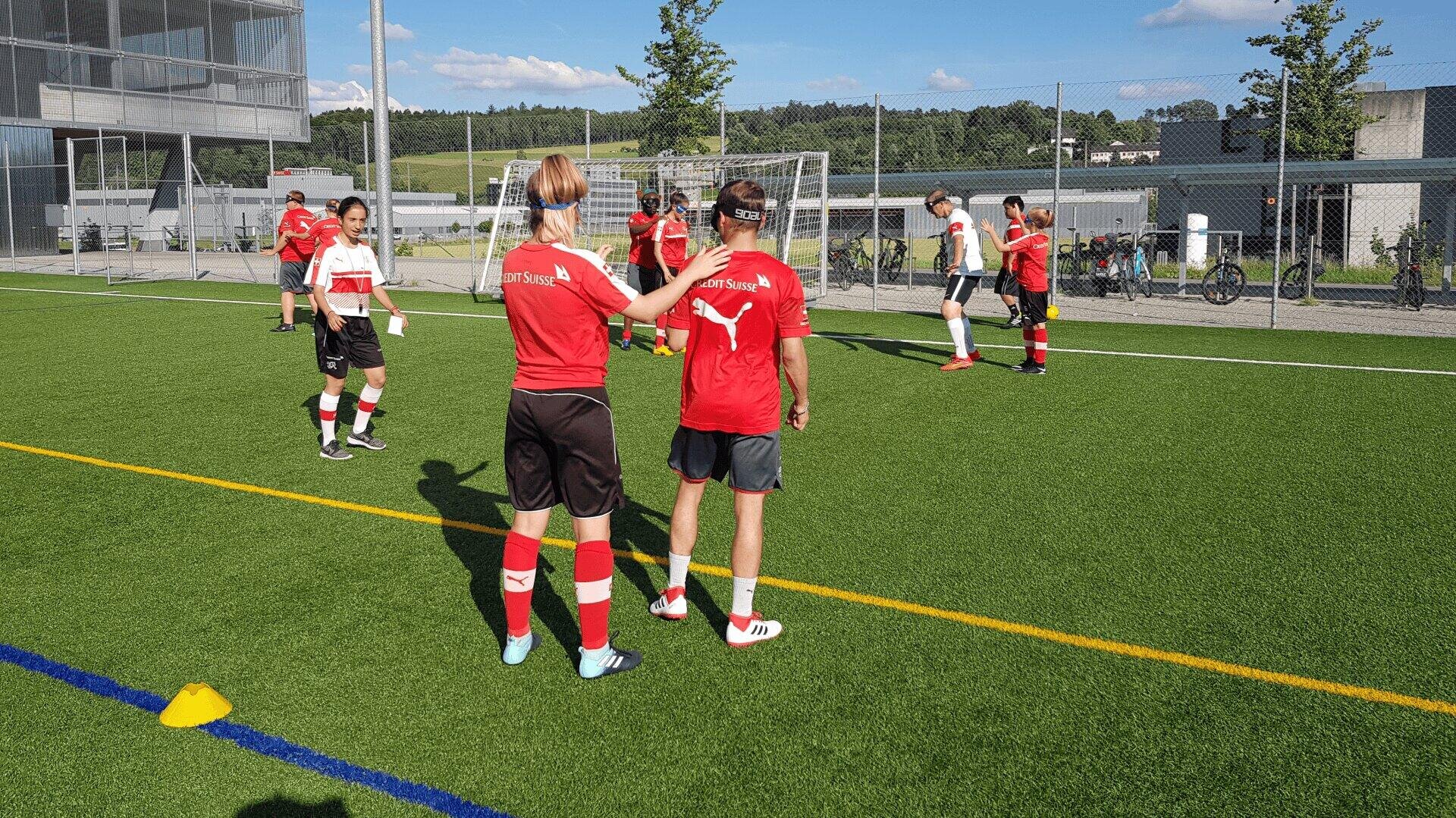 Eine Gruppe junger Fußballspieler in rot-weißen Trikots steht auf einem Kunstrasenplatz in der Nähe eines Tores und unterhält sich, wobei der Bereich durch Kegel markiert ist. Im Hintergrund sind Gebäude und Bäume unter einem sonnigen Himmel zu sehen.