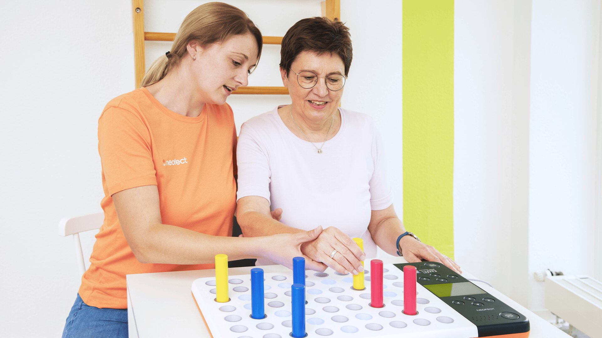 Eine jüngere Frau hilft einer älteren Frau, in einem hellen, modernen Raum farbige Stifte auf ein Brettspiel zu setzen, wahrscheinlich als Teil einer Therapie- oder Rehabilitationsmaßnahme.
