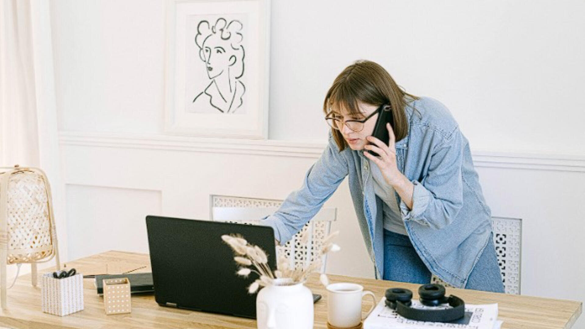 Eine Frau mit Brille telefoniert, während sie an einem hölzernen Schreibtisch mit Kaffeetasse, Notebook, Kopfhörern und Vase an einem Laptop arbeitet; an der Wand hinter ihr hängt ein minimalistisches Porträt.