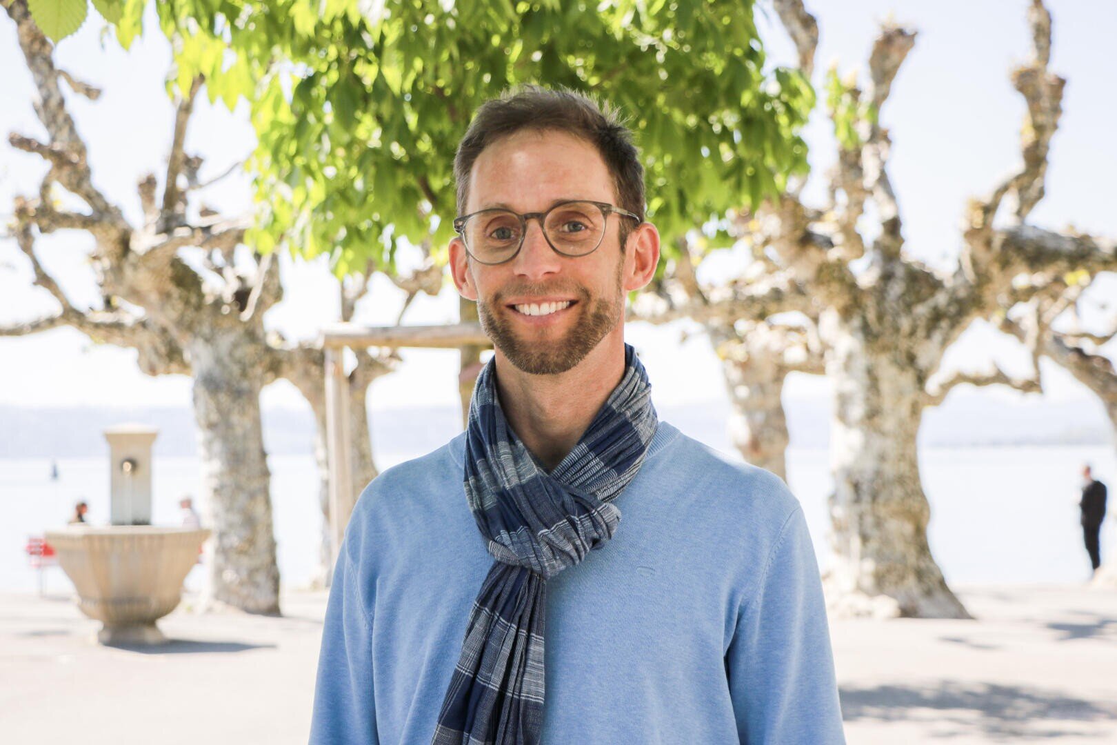 Ein lächelnder Mann mit Brille und einem blauen Schal steht draussen vor einigen Bäumen.