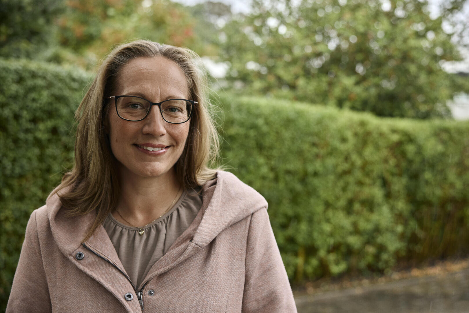 Eine Frau mit Brille und schulterlangem Haar, die einen hellrosa Mantel und ein beiges Oberteil trägt, lächelt, während sie im Freien vor einer grünen Hecke steht. Der Hintergrund ist mit Bäumen und bedecktem Himmel leicht unscharf.
