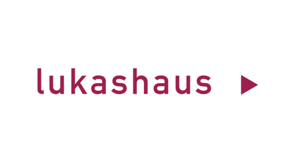 Das Logo von "lukashaus" ist in rot geschrieben und endet mit einem roten Dreieck, das nach rechts zeigt.