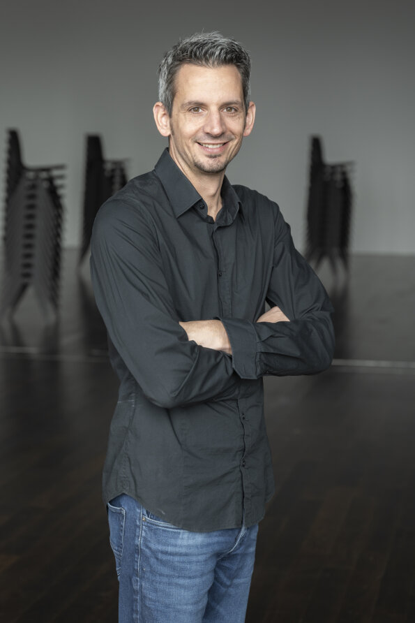Ein Mann mit kurzen grauen Haaren, der ein dunkles Button-up-Hemd und blaue Jeans trägt, steht mit verschränkten Armen in einem Haus und lächelt in die Kamera. Im unscharfen Hintergrund sind gestapelte Stühle zu sehen.