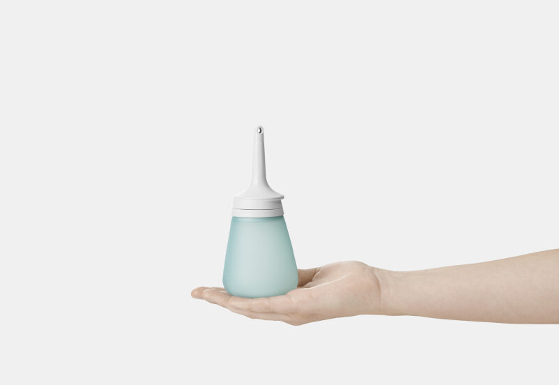 Eine Hand hält eine blaue Quetschflasche mit einer langen weißen Tülle, die üblicherweise als Nasenspülflasche verwendet wird, vor einem schlichten weißen Hintergrund.