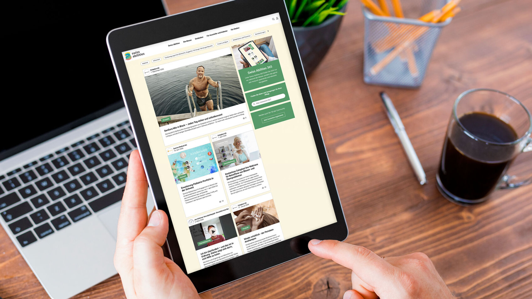 Eine Person, die ein Tablet in der Hand hält, auf dem eine Website mit Artikeln zu Gesundheit und Wellness angezeigt wird; auf einem Holztisch mit einem Laptop, einem Stifthalter und einer Tasse schwarzem Kaffee.