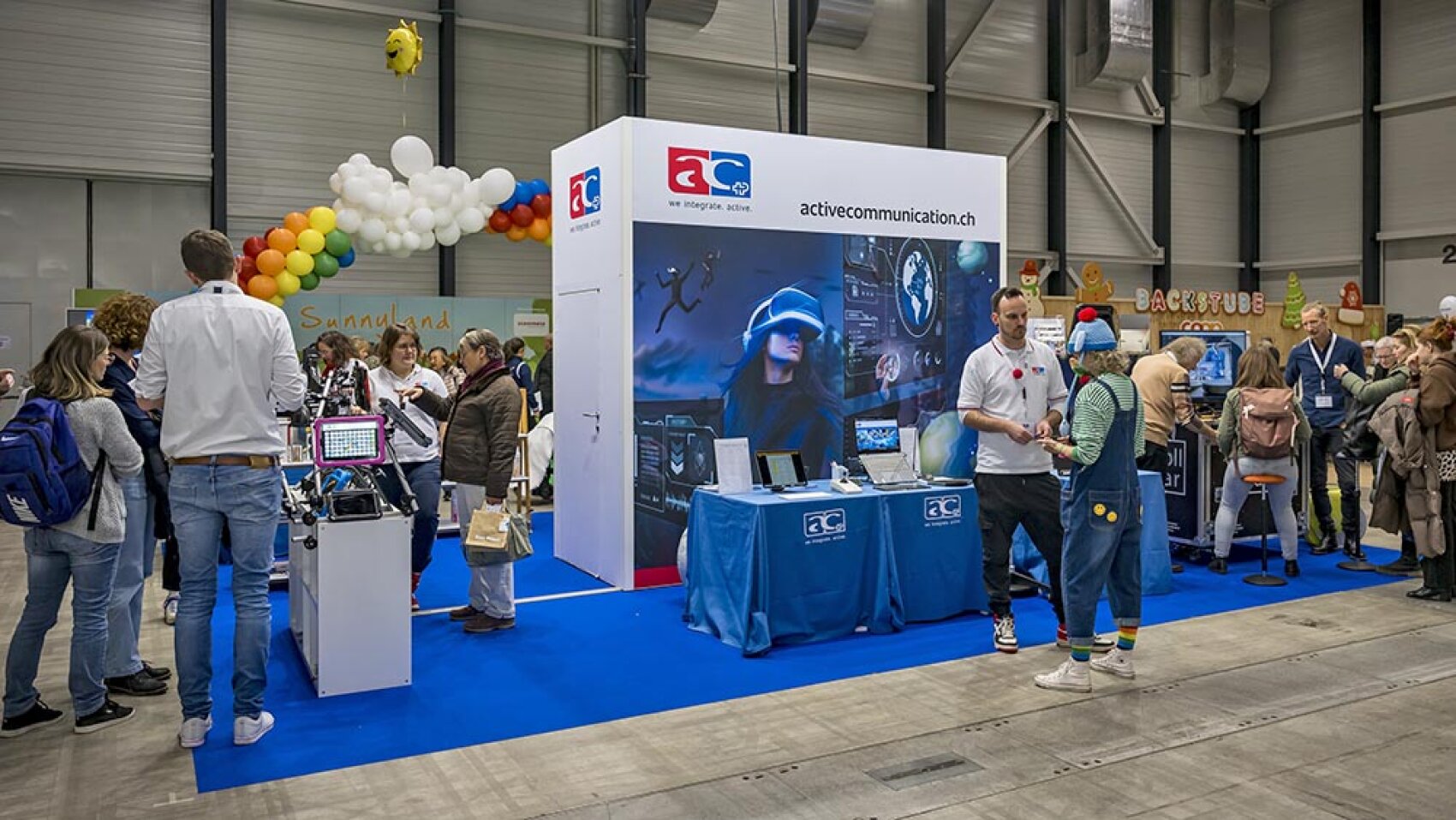 Auf einem belebten Messestand mit blauen Teppichen werden Technologieprodukte ausgestellt. Menschen interagieren mit Geräten und Mitarbeitern, während bunte Ballons und Banner den Bereich schmücken. Ein großes Poster zeigt eine Person, die virtuelle Realität nutzt.